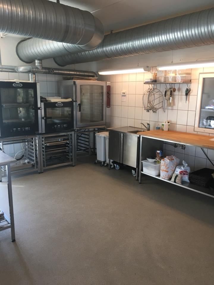 Et stort kommercielt køkken med ovne, hætte, skabe, håndklæder, tøj og flere hylder med ting. En stor metallisk luftkanal