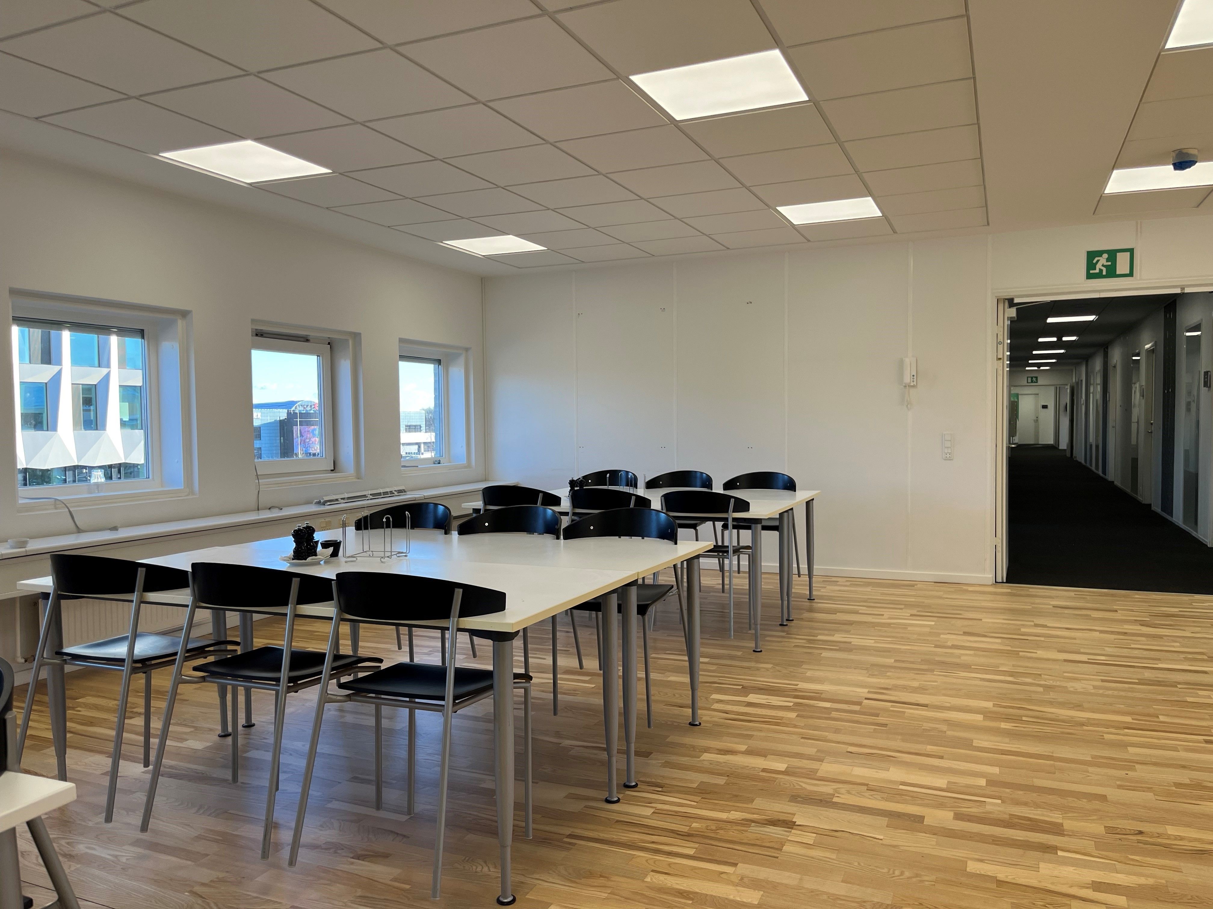 Kontorlokaler med hvide vægge, lyse loftslampe og trægulve, flere runde borde med stole og to døre i enden af lokalet