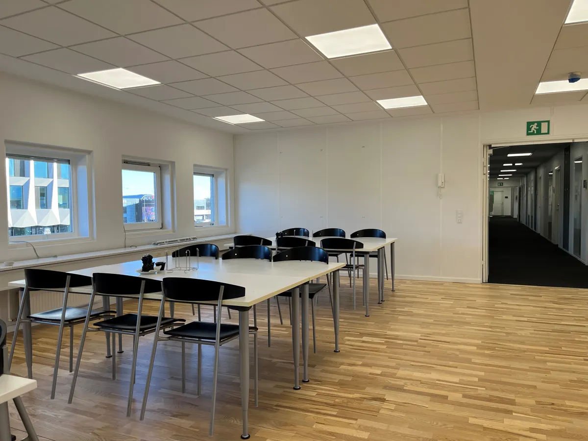 Kontorlokaler med hvide vægge, lyse loftslampe og trægulve, flere runde borde med stole og to døre i enden af lokalet