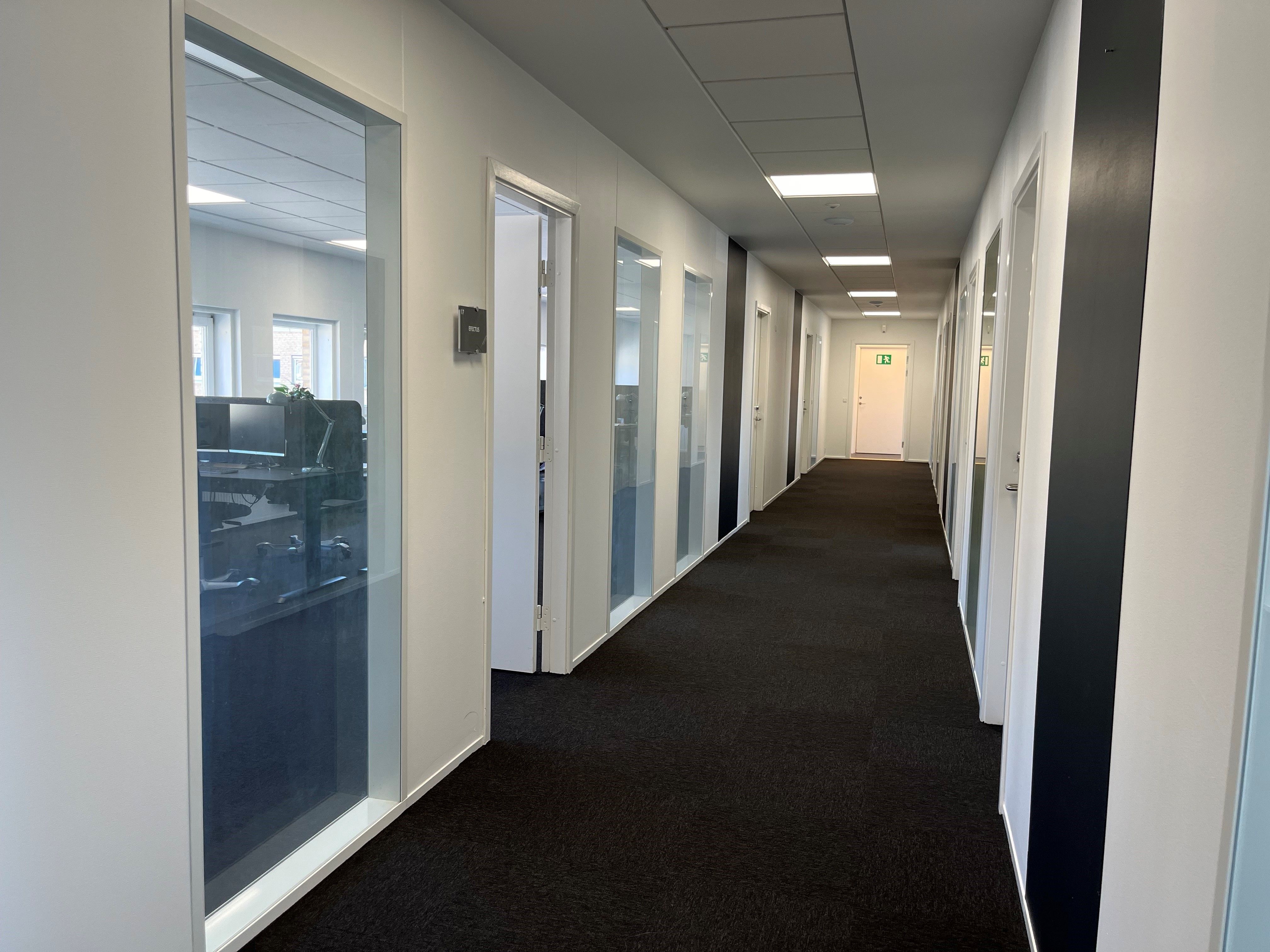 Kort og smal kontorhvile med hvide vægge og sortgule døre med håndtag, glasdøre, mørke gulvbelægning, lyse loftsbærere og