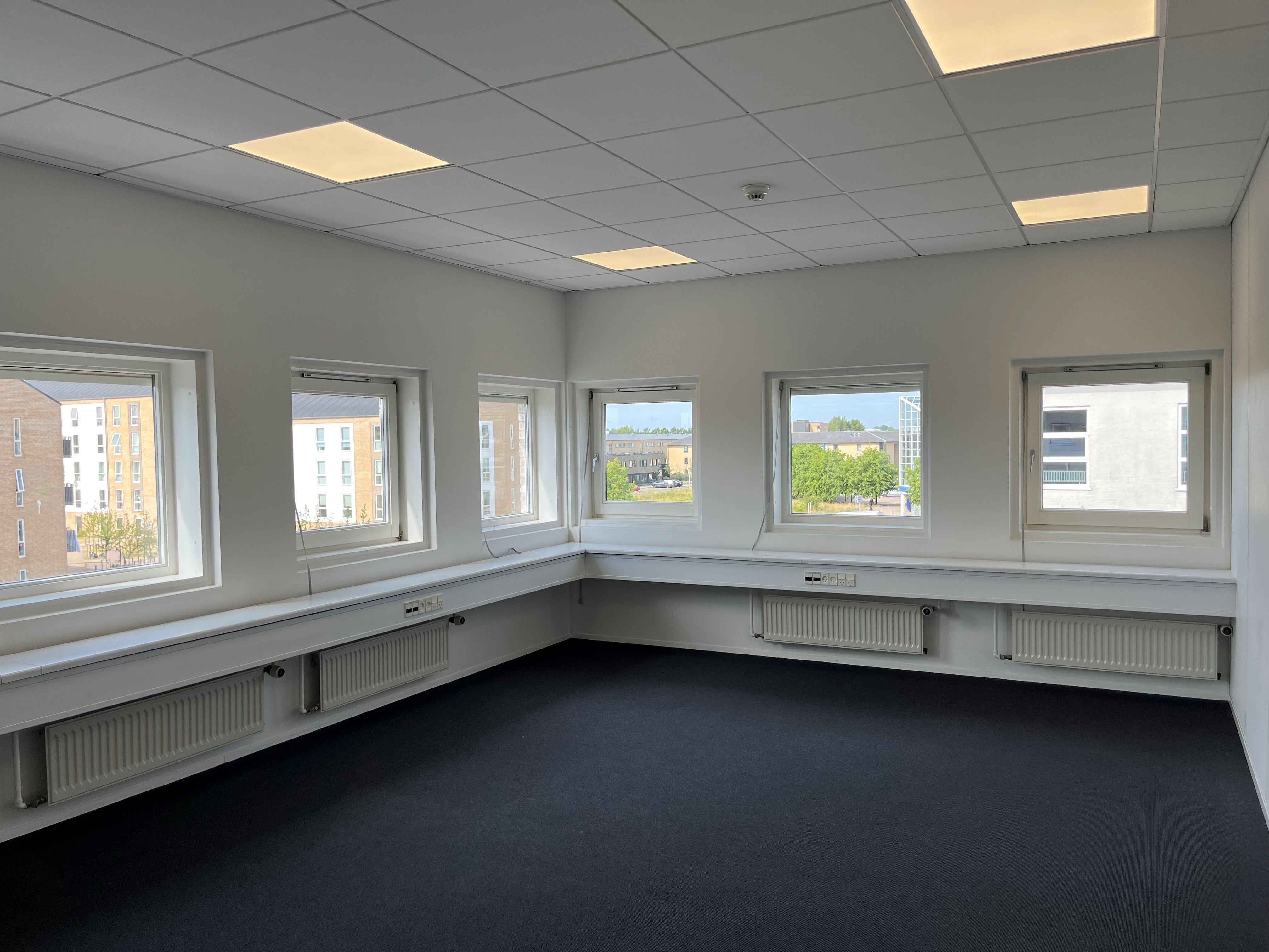 Modernt kontor med hvide vægge, lyse loftspaneler, lyse vinduer, radiatorer, og sort gulvbelægning