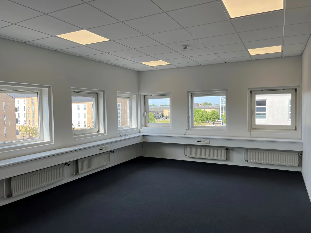 Modernt kontor med hvide vægge, lyse loftspaneler, lyse vinduer, radiatorer, og sort gulvbelægning