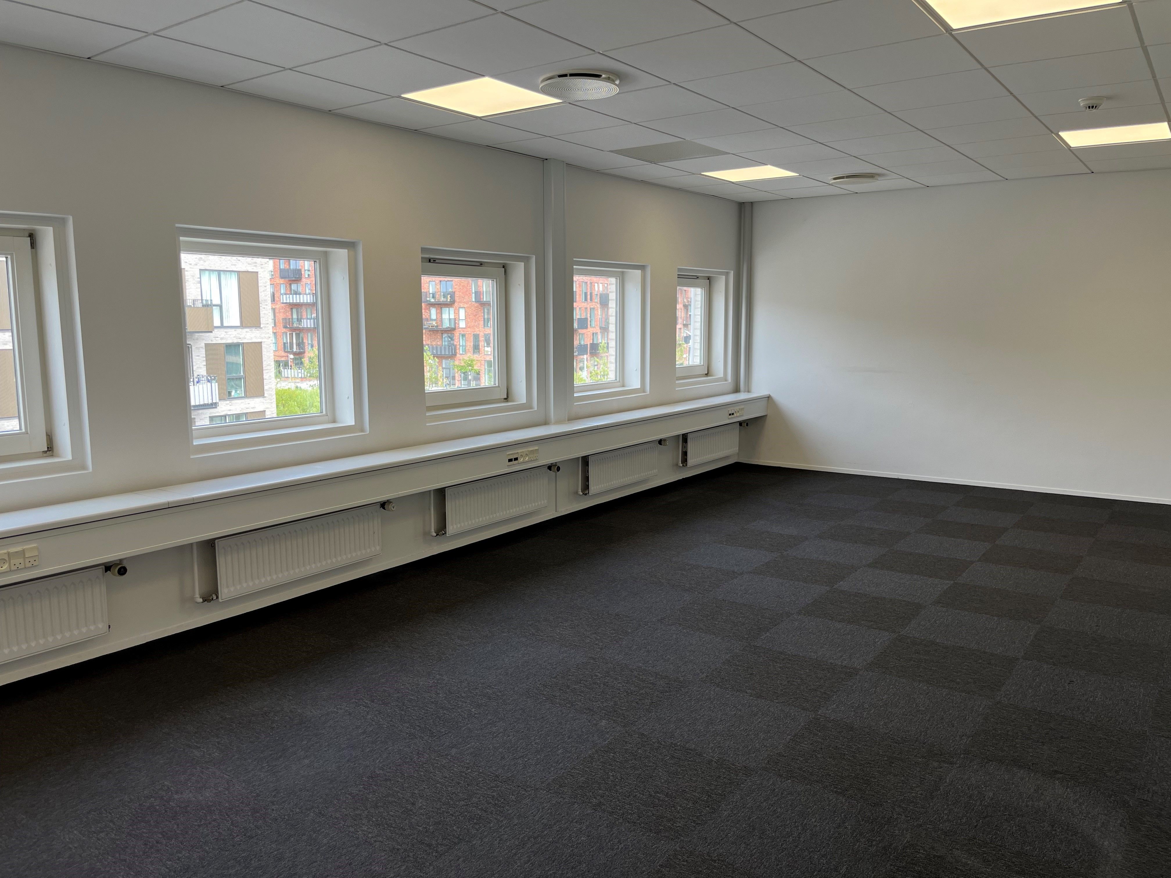 En stor kontorplads med fire vinduer og lyse gulvbelægning og hvide vægge samt en række radiatorer under vinduet