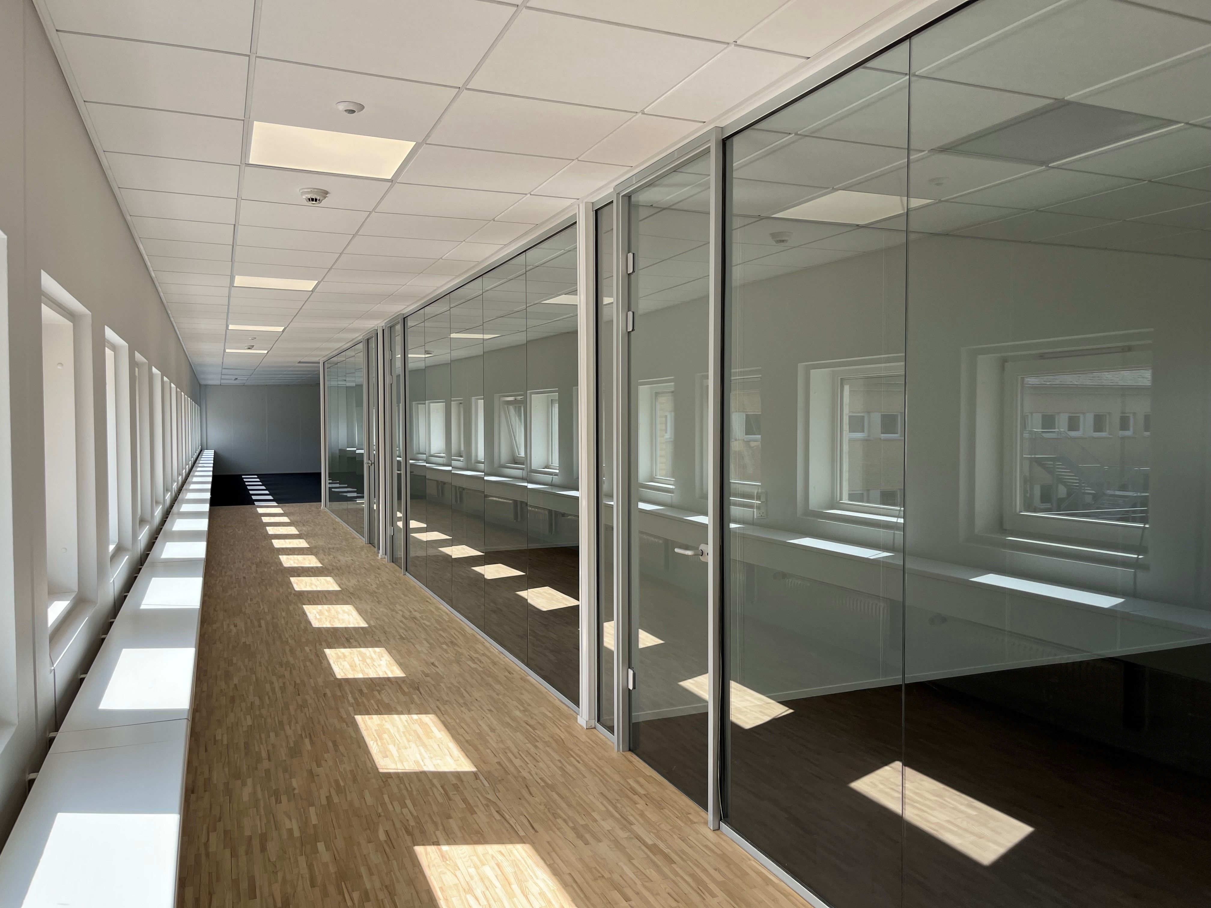 Hvidt lokale med glasdøre og gulv af træ. Vægge og loft er hvide og lyse. Solskin trænger ind gennem vinduerne og kaster