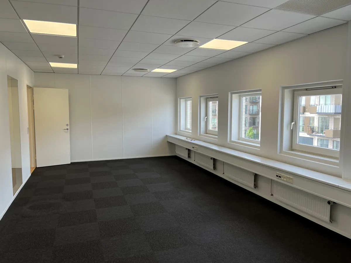 En tom, lys og moderne kontor med hvide vægge, sort gulv og fire vinduer med en udsigt til en bygning med balkoner.