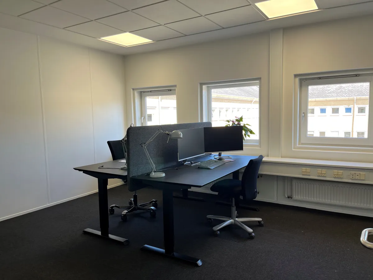 Kontor med to skriveborde, hver med en lysegrå stol, en lampeskærm og to skærme, samt en blomst på vindueskarme