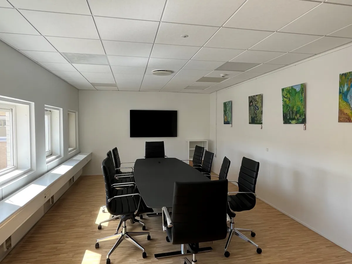 Mødelokale med moderne møbler og en stor tv-skærm samt malerier på væggen og vinduer med udsigt til gaden