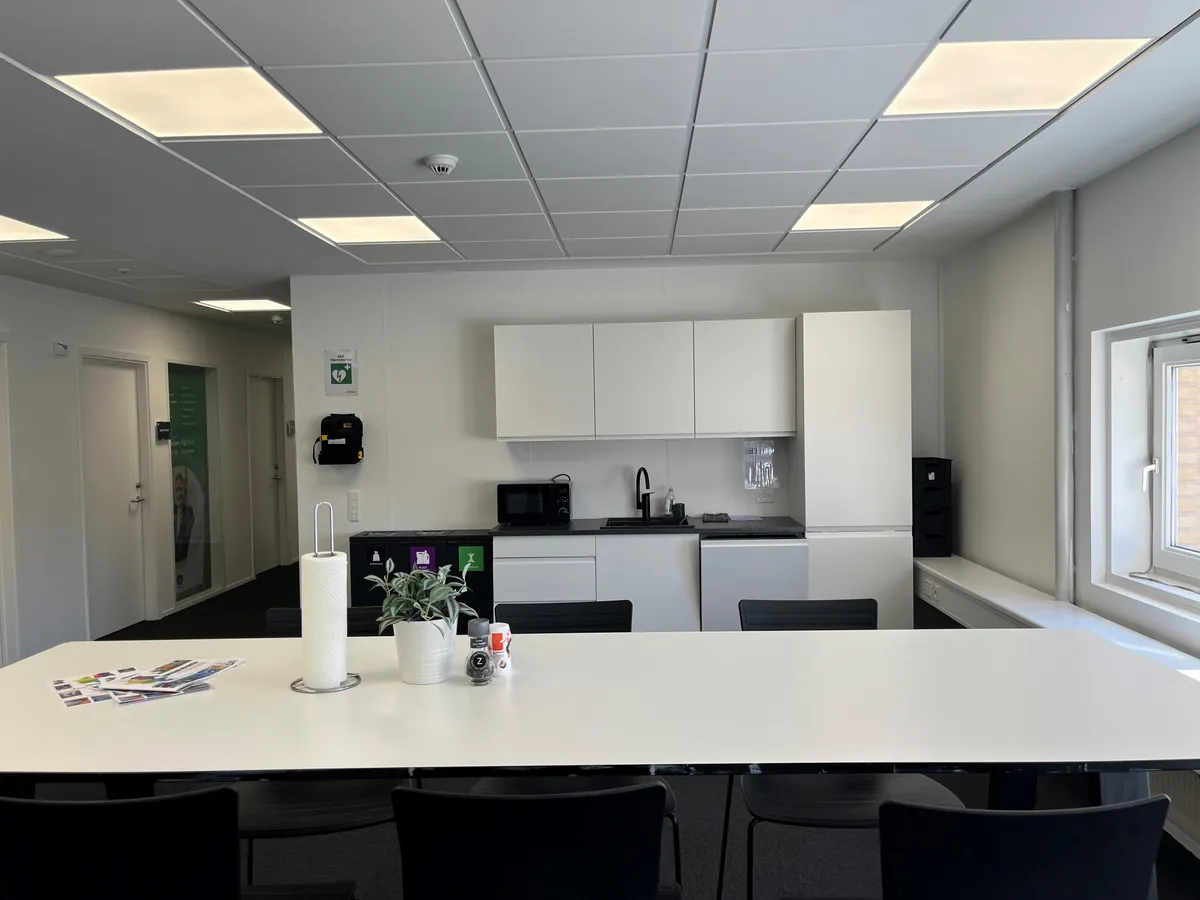 Mødelokale med hvide vægge og loft, fuldt udstyret køkken, og tomme møbler og skrivebord