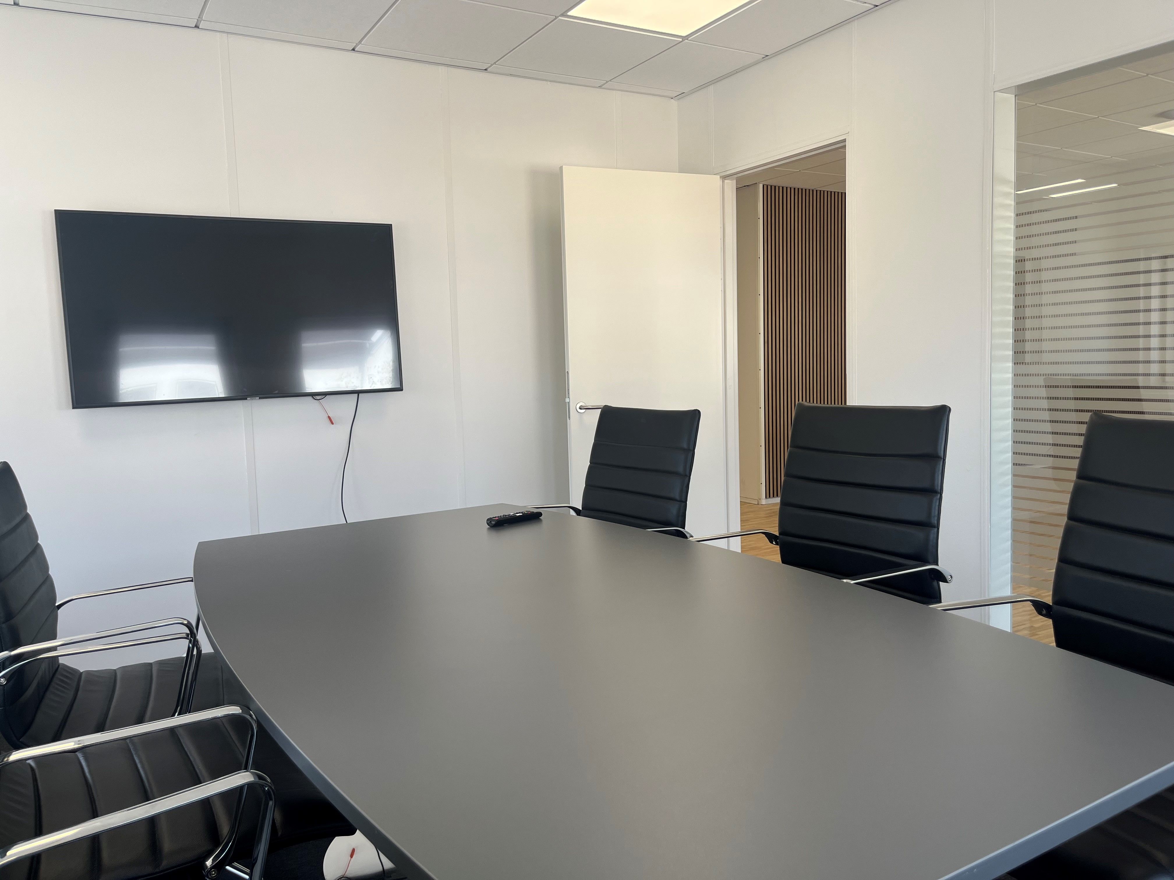 En mødelokale med en stor fladskærms-tv og fire sorte læderstole omkring et langt bord med en fjernbetjening
