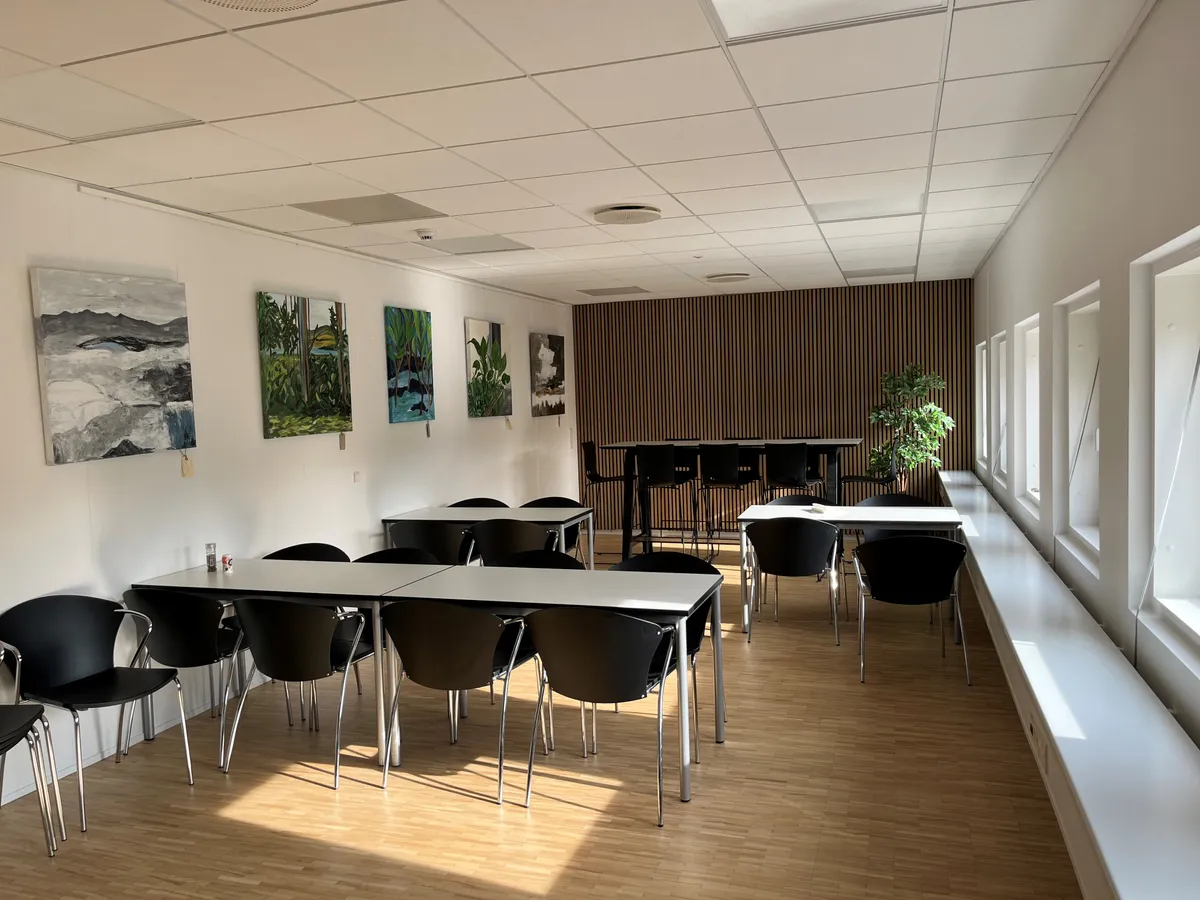 Moderne kontorlokaler med to lange borde, fire små borde, stolpe, kunstværker og vinduer på den ene side af rummet