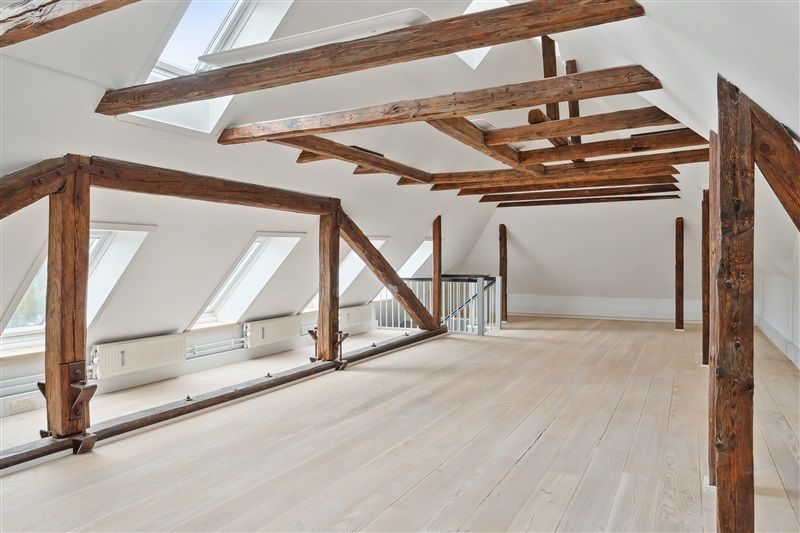 Loftsrum med højt loft, vinduer og træbjælker samt hvide vægge og gulv