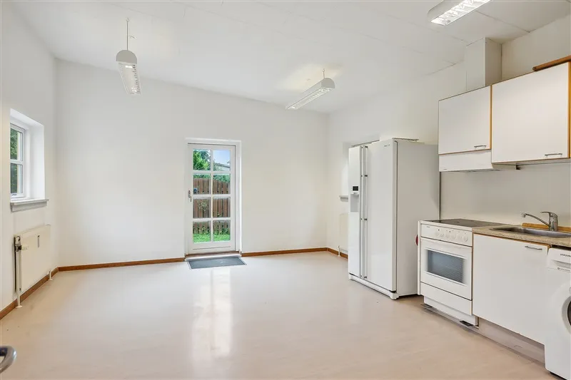 En moderne køkken med hvide vægge, hvidt køkkenskab, køleskab, komfur, vask og en dør til terrassen