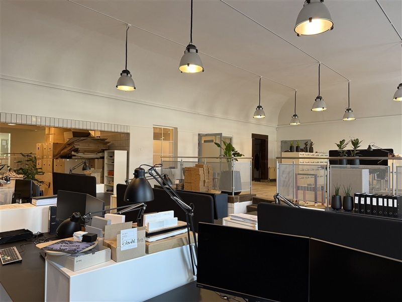 Kontorlokaler med skrivebord, monitor, skrivebordslampe, og flere skriveborde med computere og planter