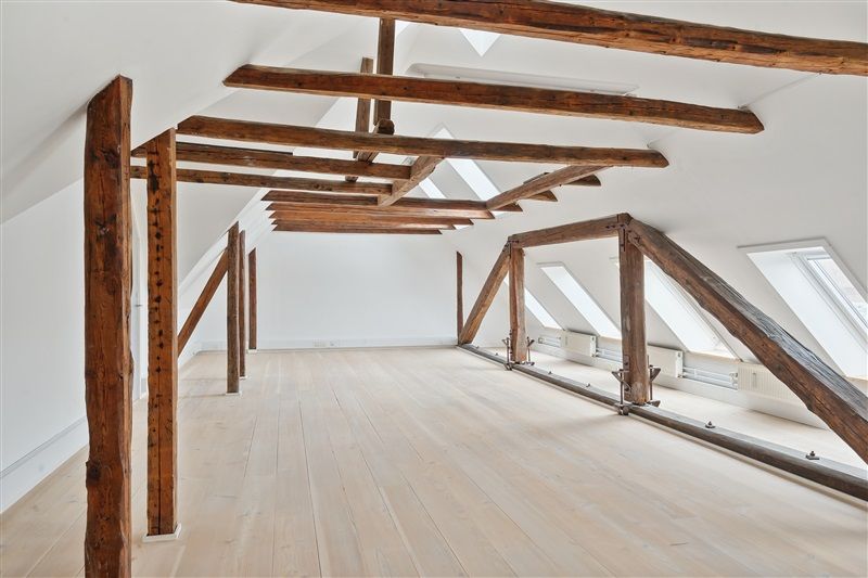 Loftsrum med lyse træbjælker, hvide vægge og lyse gulv samt vinduer og radiatorer på væggen