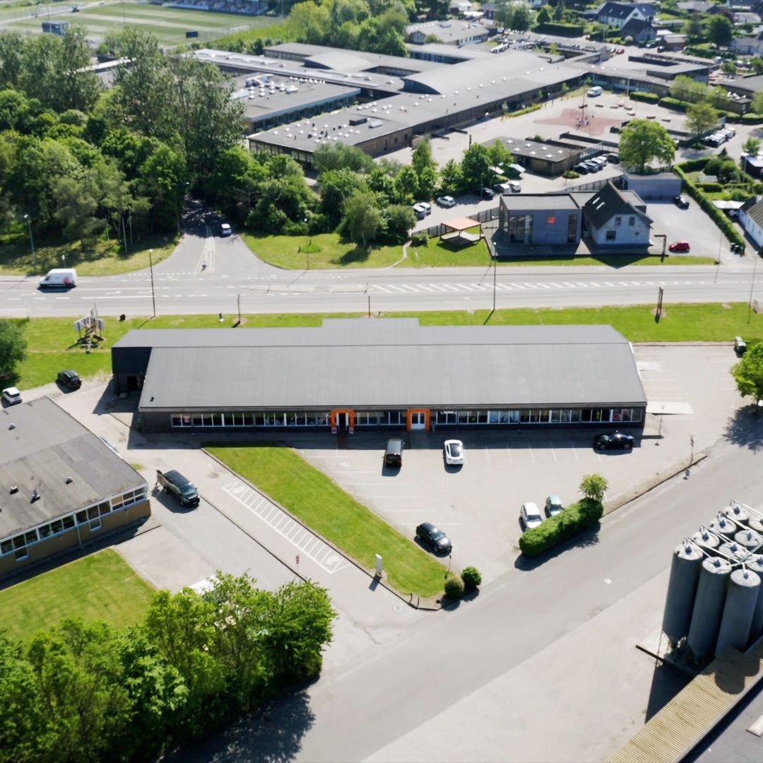 Butikslokaler med parkeringsplads og grønne områder, forskellige biler parkeret, udendørs skulptur og grønne træer