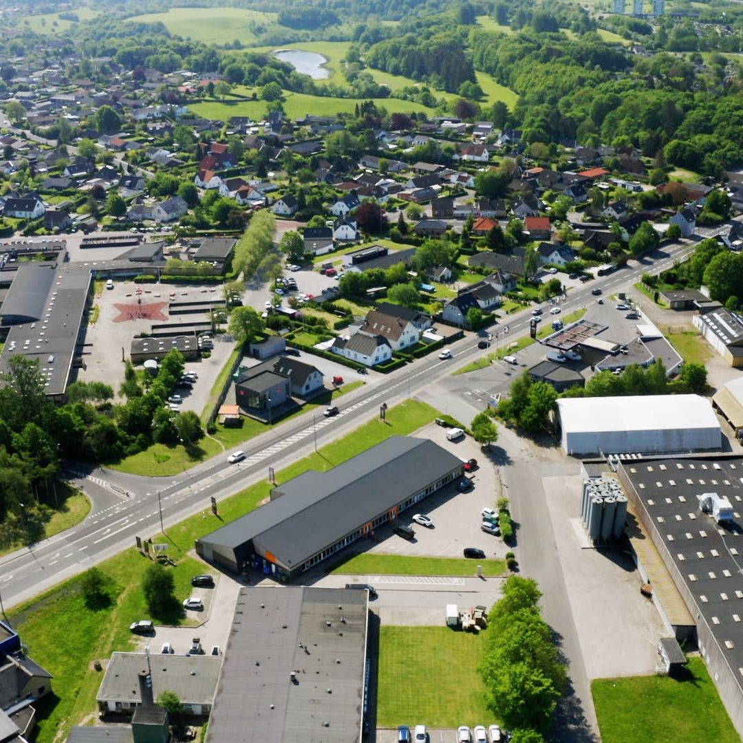 Luftfoto af et byområde med bygninger, veje og parkeringspladser, fuld af grønne træer og buske