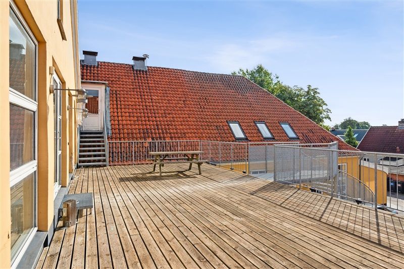 Træterrasse med udsyn til bygninger med tegltag og træranker, samt et træbord og en metalkælder
