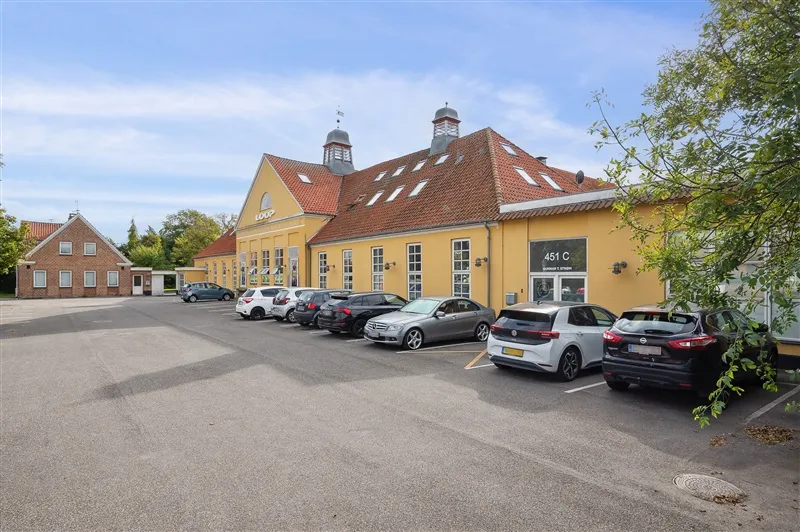 Parkeringsplads foran bygning med skiltet LODP og nummer 451 C, med flere biler parkeret og en stor trægren