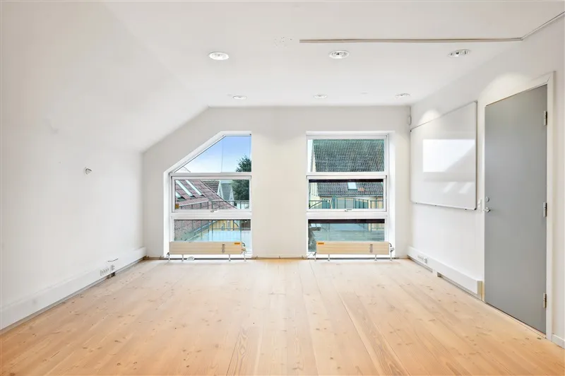 En tom kontorlokal med lyse trægulve og hvide vægge samt to store vinduer med glaspaneler