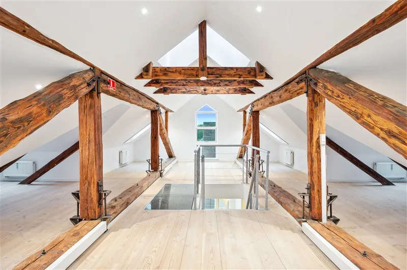 Loftsrum med højloft og lyse loftsbjælker i træ samt glasvinduer og trægulve