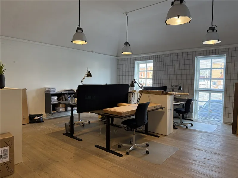 Kontor med to skriveborde og stol, vinduer med solblinde, to lyskroner og to plantekrukker samt en hvid stol og et gulv med