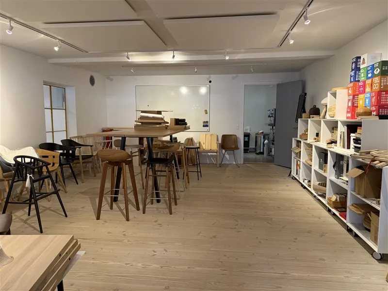 Hvidt rum med lyse trægulve og højt til loftet. Bord og stole samt et lysebord. Skabe med hylder og mange bokse.