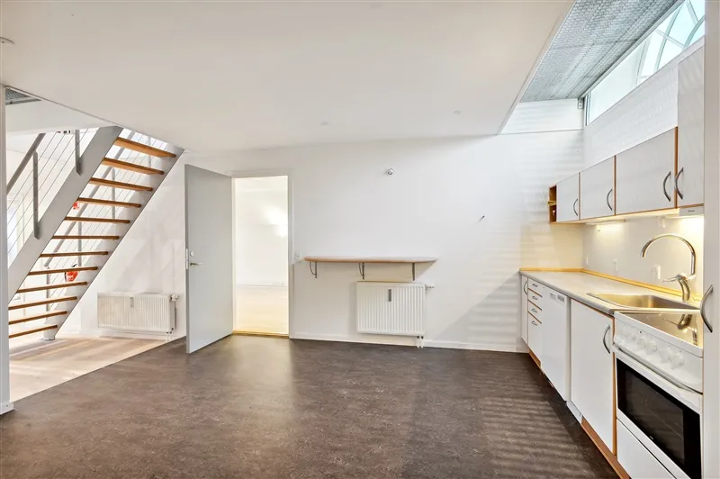 Rent og pænt køkken med hvide vægge og gulv, hvidt køkken med lysegrå skabe, en ovn, og et vinduesfelt med lys fra vindue i