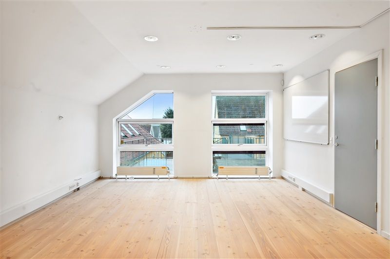 To store vinduer med udsigt til gårdhave og grønne træer samt en hvid væg med en whiteboard og en lukket dør