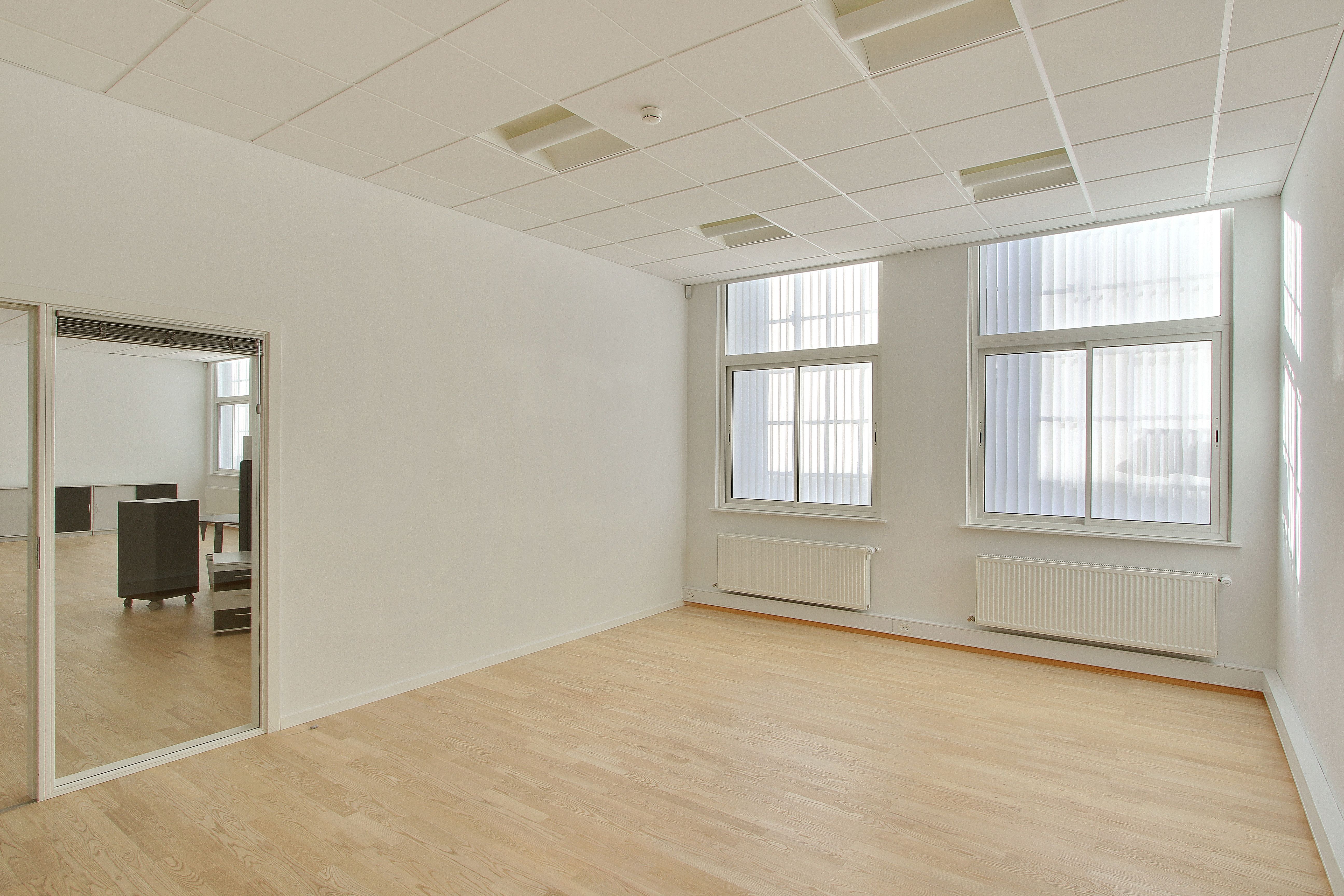 Et moderne kontor med hvide vægge og gulve samt store vinduer med jalousier. Et enkelt højt spejl og en lyse radiatorer.