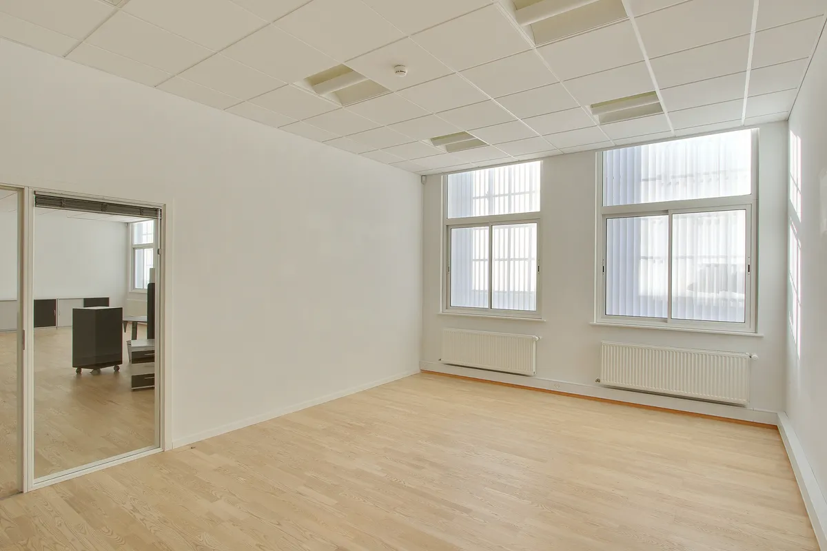 Et moderne kontor med hvide vægge og gulve samt store vinduer med jalousier. Et enkelt højt spejl og en lyse radiatorer.