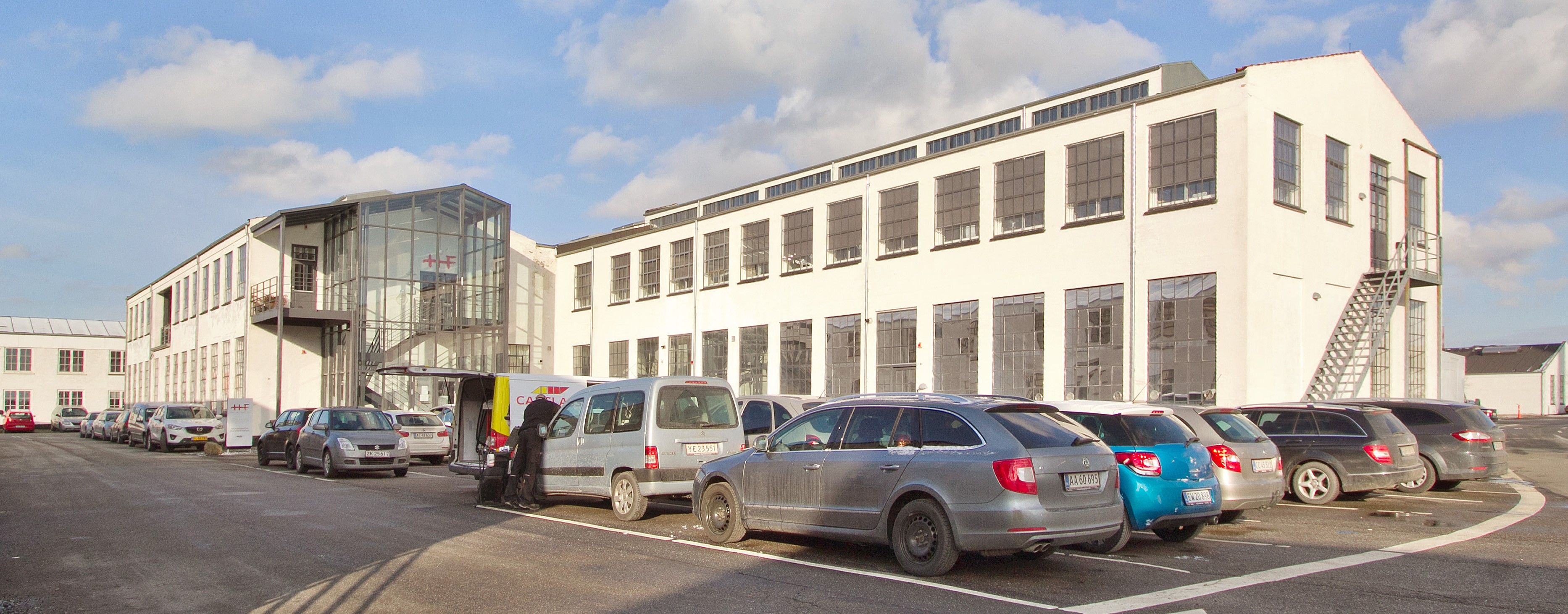 Parkerede biler udenfor et hvidt bygningskompleks med flere vinduer, et højt stige og en røgfrastøjningsanlæg.