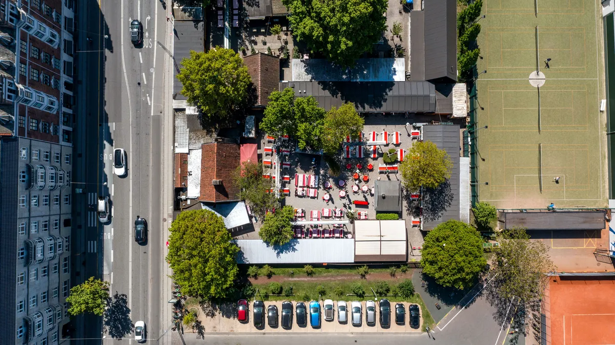 Et ølhave med røde og hvide stole og borde samt parkerede biler langs siden af en gade med tennisbaner og huse