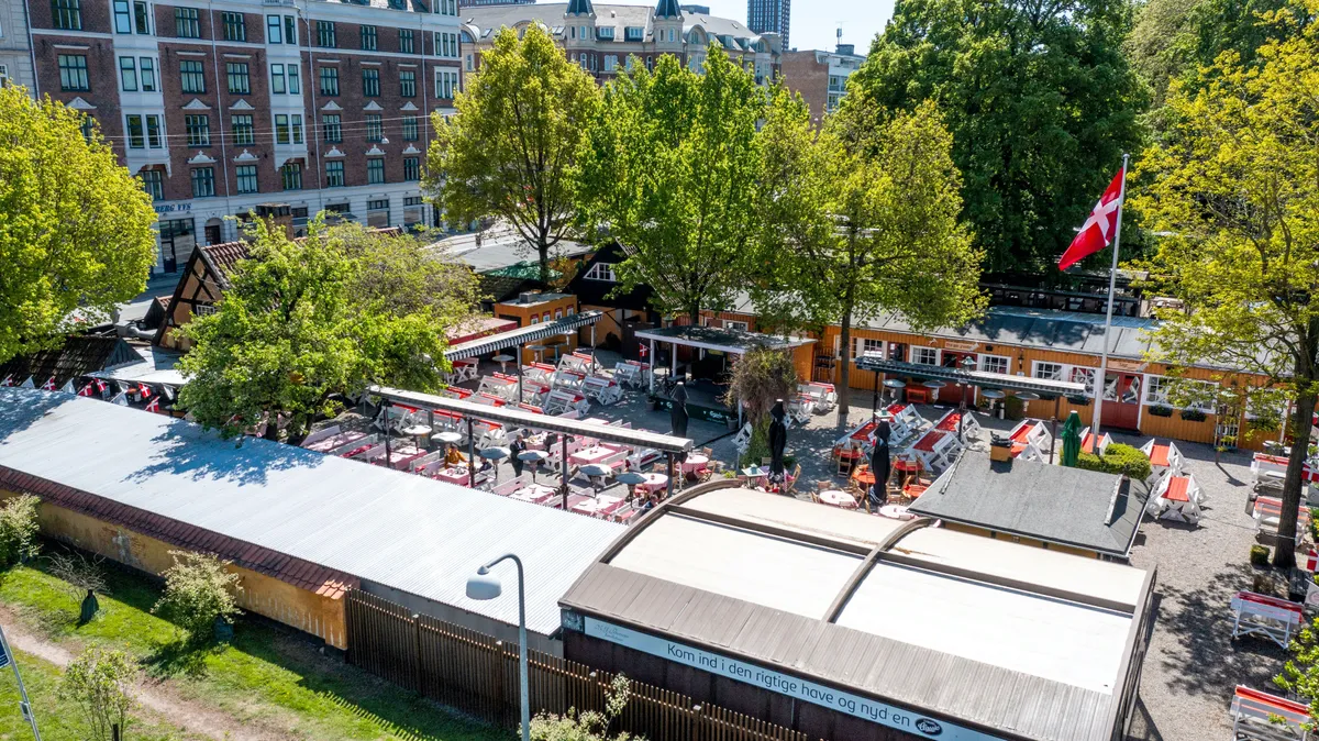 En ølhave med træhytter og flag, små borde og stole i haven og store bygninger i baggrunden