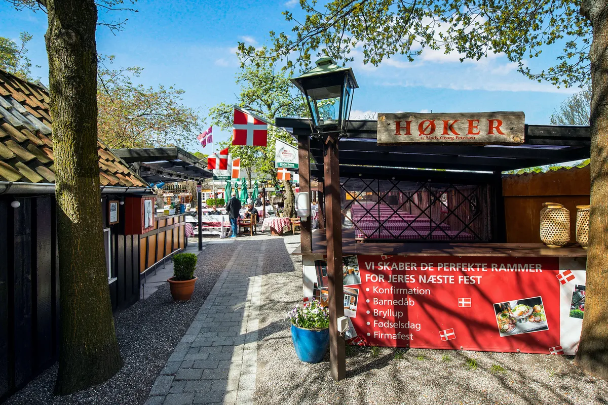 En ølstand med sammenklappelige bænke og bord under søjler og lysekroner, med skilte og flag og små huse i baggrunden