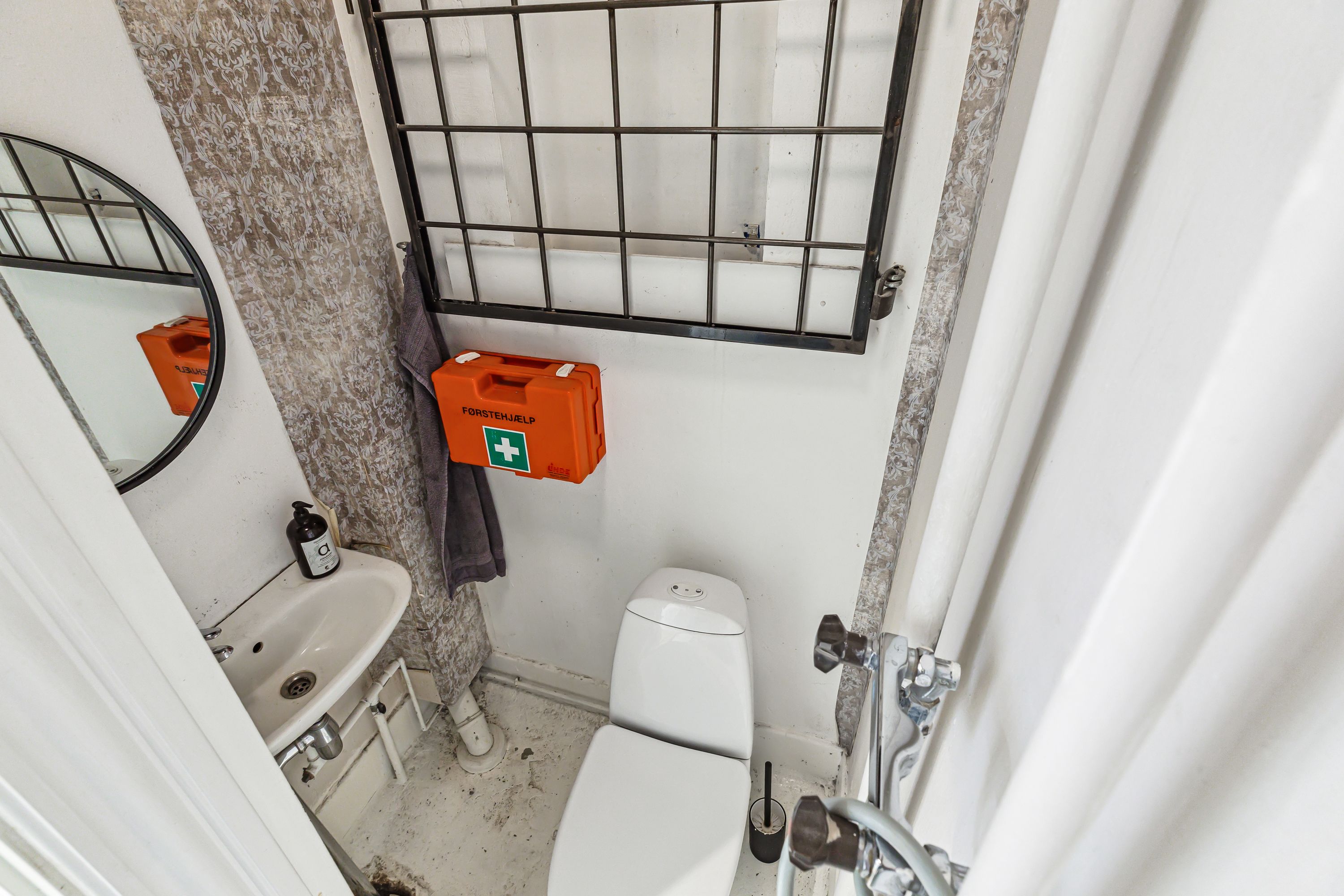 Et lille badeværelse med en toilet, håndvask og et førstehjælpskasse på væggen og et spejl med en anden førstehjælpskasse i
