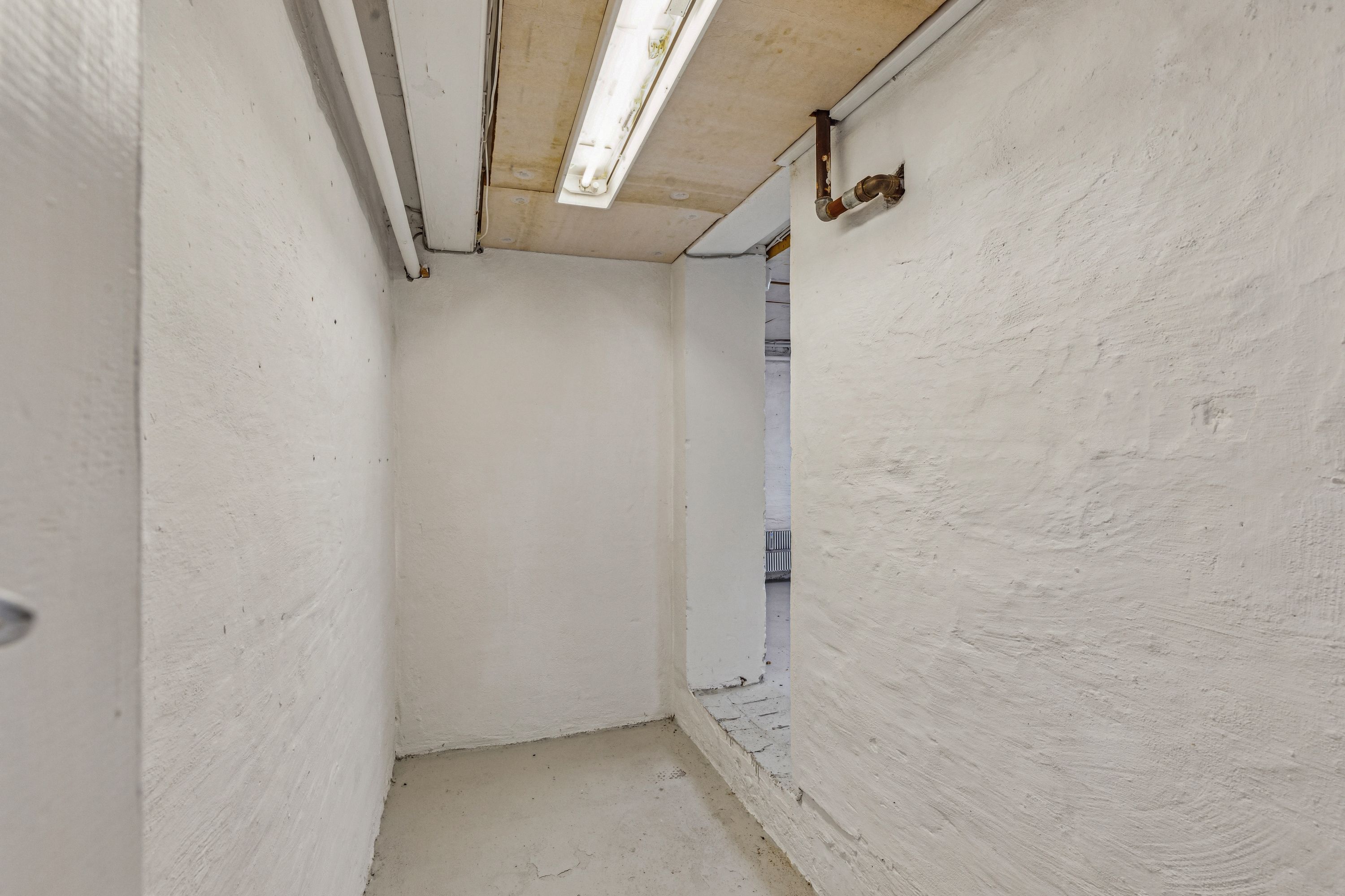 En trang, ubelyst gang med hvide vægge og gulv samt en åbning til næste rum med rør og røgskafte