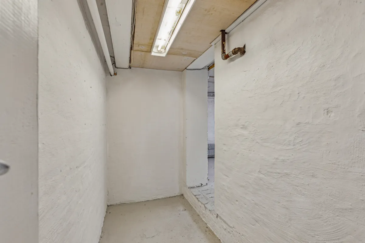 En trang, ubelyst gang med hvide vægge og gulv samt en åbning til næste rum med rør og røgskafte