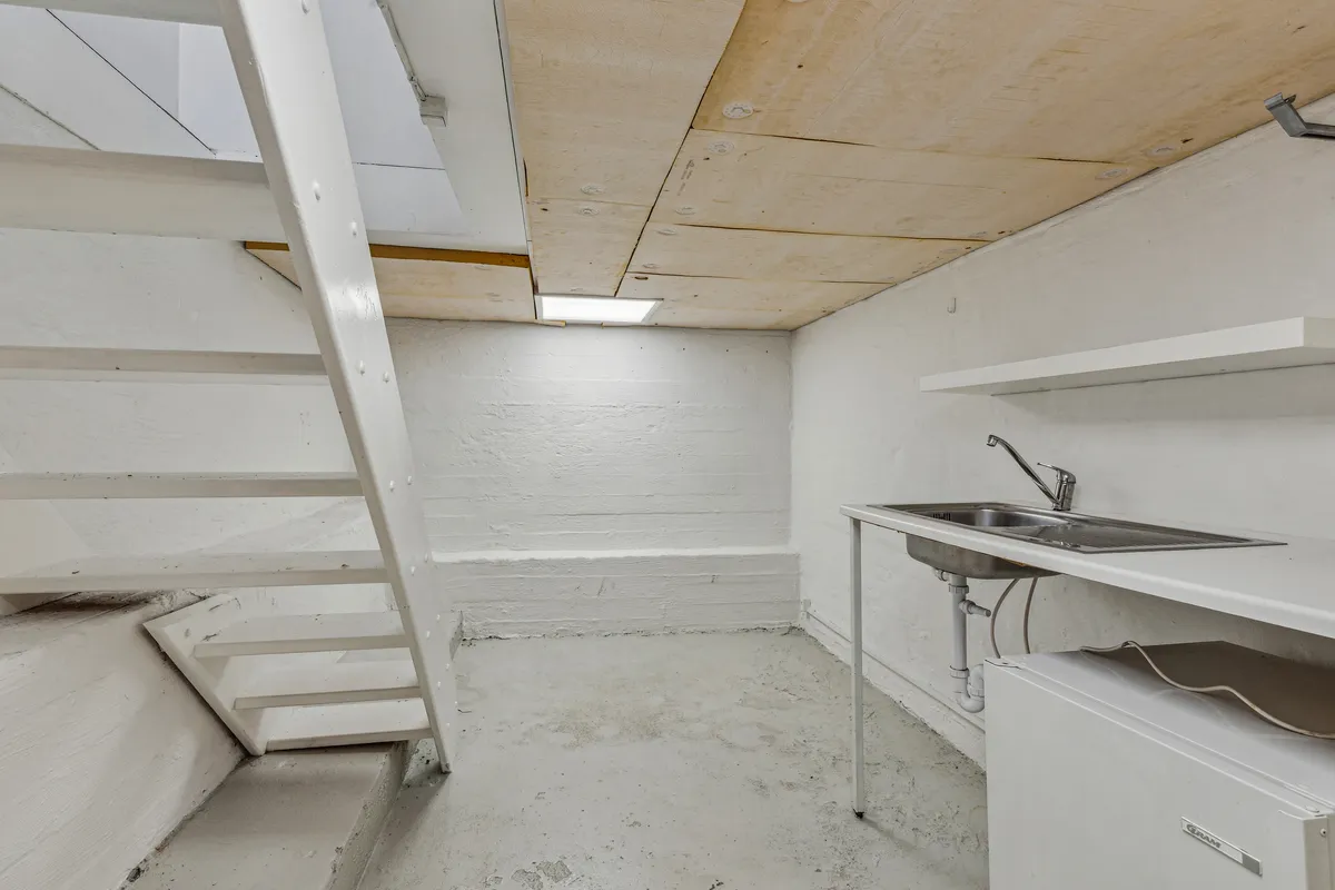 Et lille værelse med hvide vægge og loft samt en metaltrappe og et køkken med en skraldespand og en vandhane