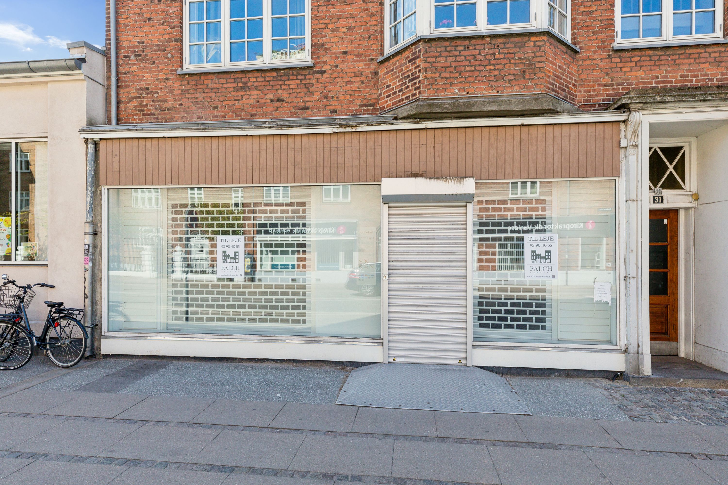 To cykler parkeret ved glasvinduer i en bygning med reklameskilte, hvor det står "TIL LEJE" og "FALCH" samt en nummerplade