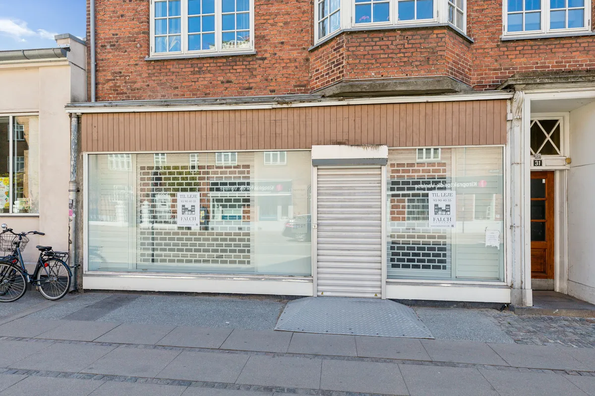 To cykler parkeret ved glasvinduer i en bygning med reklameskilte, hvor det står "TIL LEJE" og "FALCH" samt en nummerplade