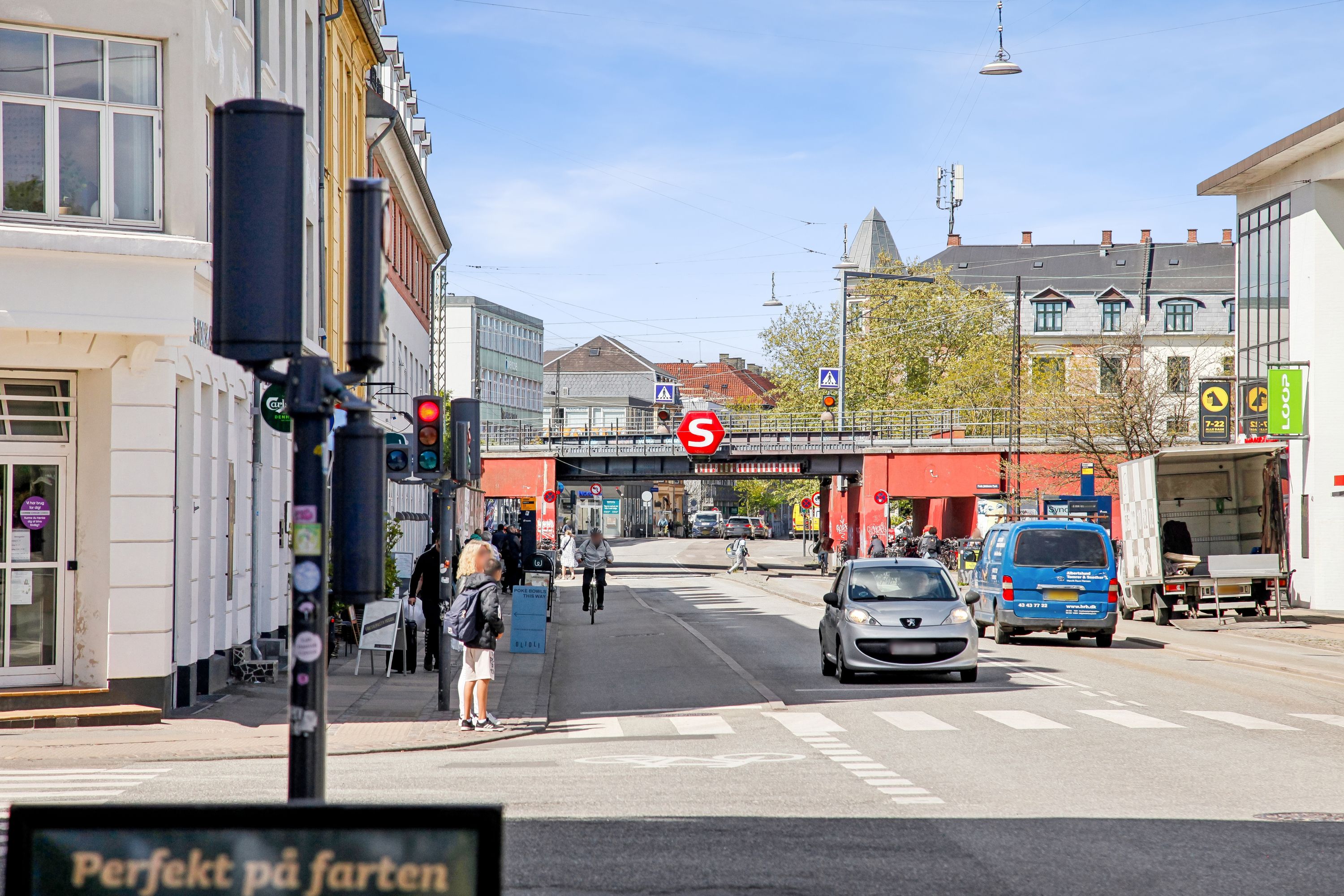 En bil kører på en gade med stopskilt og biler og folk i baggrunden