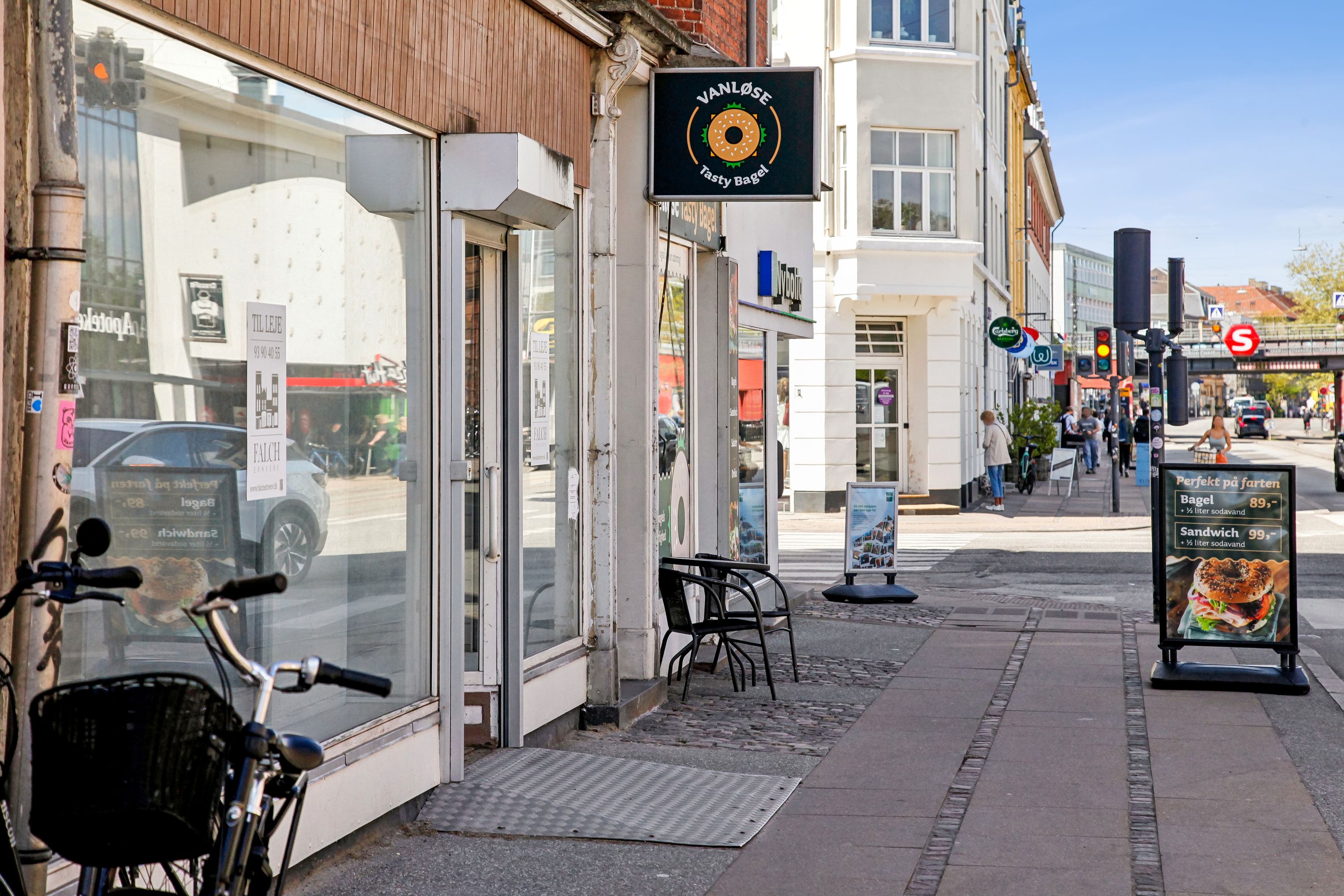 Vanløse Tasty Bagel-køkken med udendørsborde og menukort. Bygning med store ruder og biler i refleksionen. På fortovet står