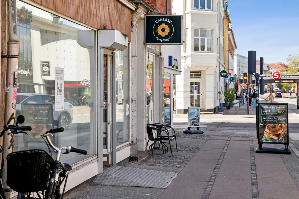 Vanløse Tasty Bagel-køkken med udendørsborde og menukort. Bygning med store ruder og biler i refleksionen. På fortovet står