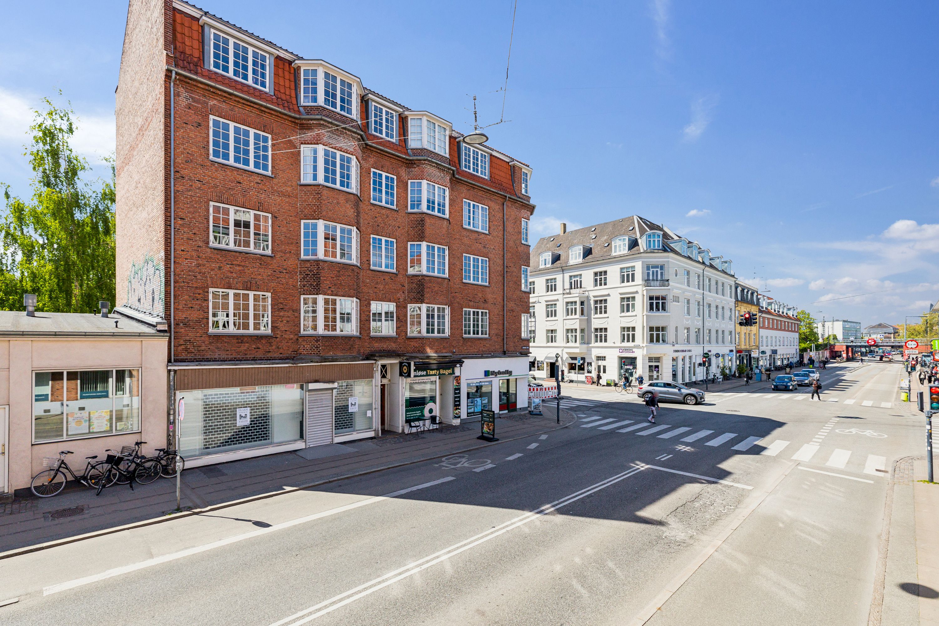 En tom og bred gade med krydsende fodgængere og biler, et rødt lys, tre cykler parkeret og nogle bygninger med vinduer