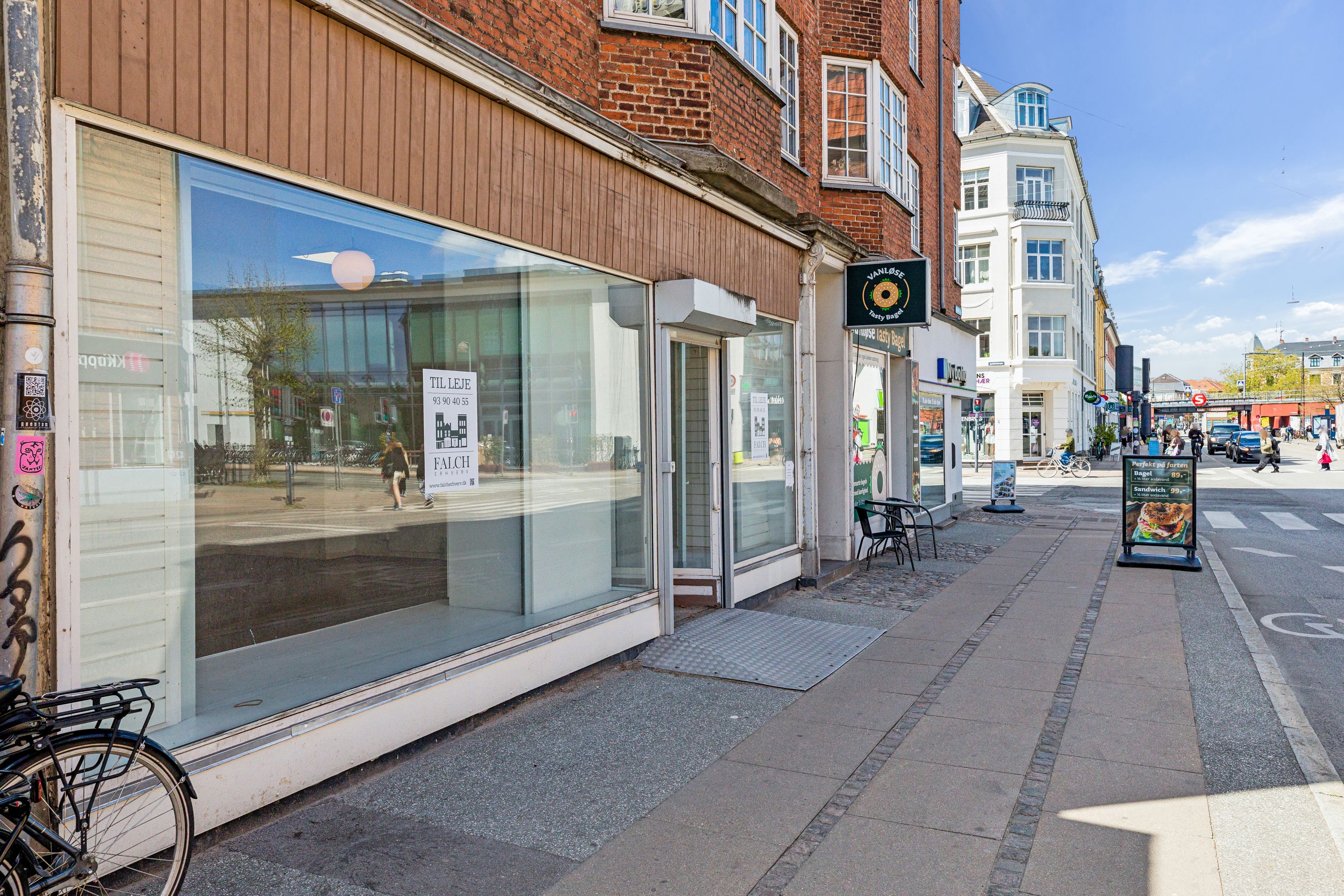 Butik med store ruder og skilte med teksten Vanløse Bakery. Der er en stol med bord og reklameskilt for sandwich og andre