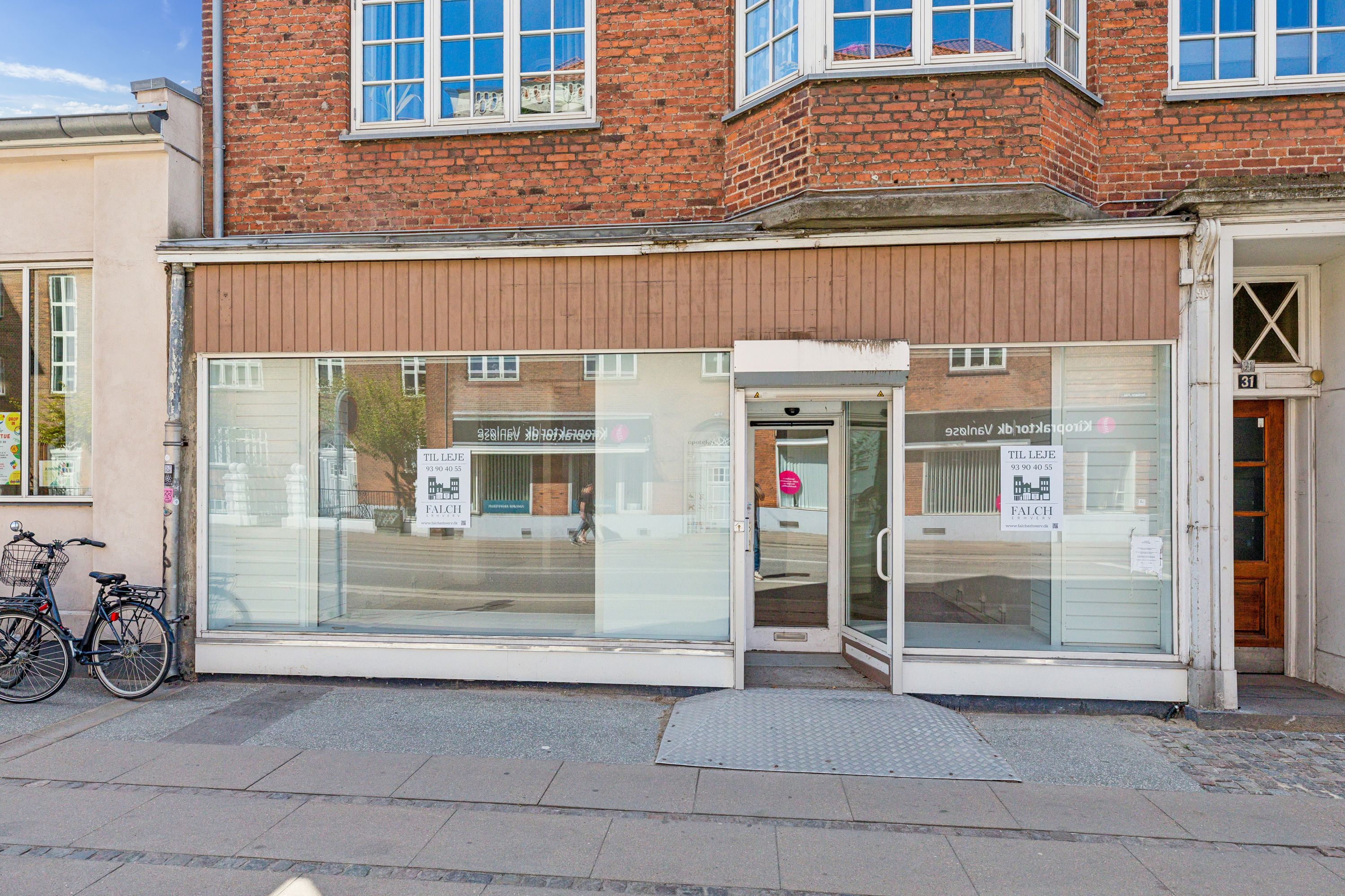 Detailhandelslokaler med glasvinduer og skilte til leje i Vanløse, med cykler parkeret foran bygningen.