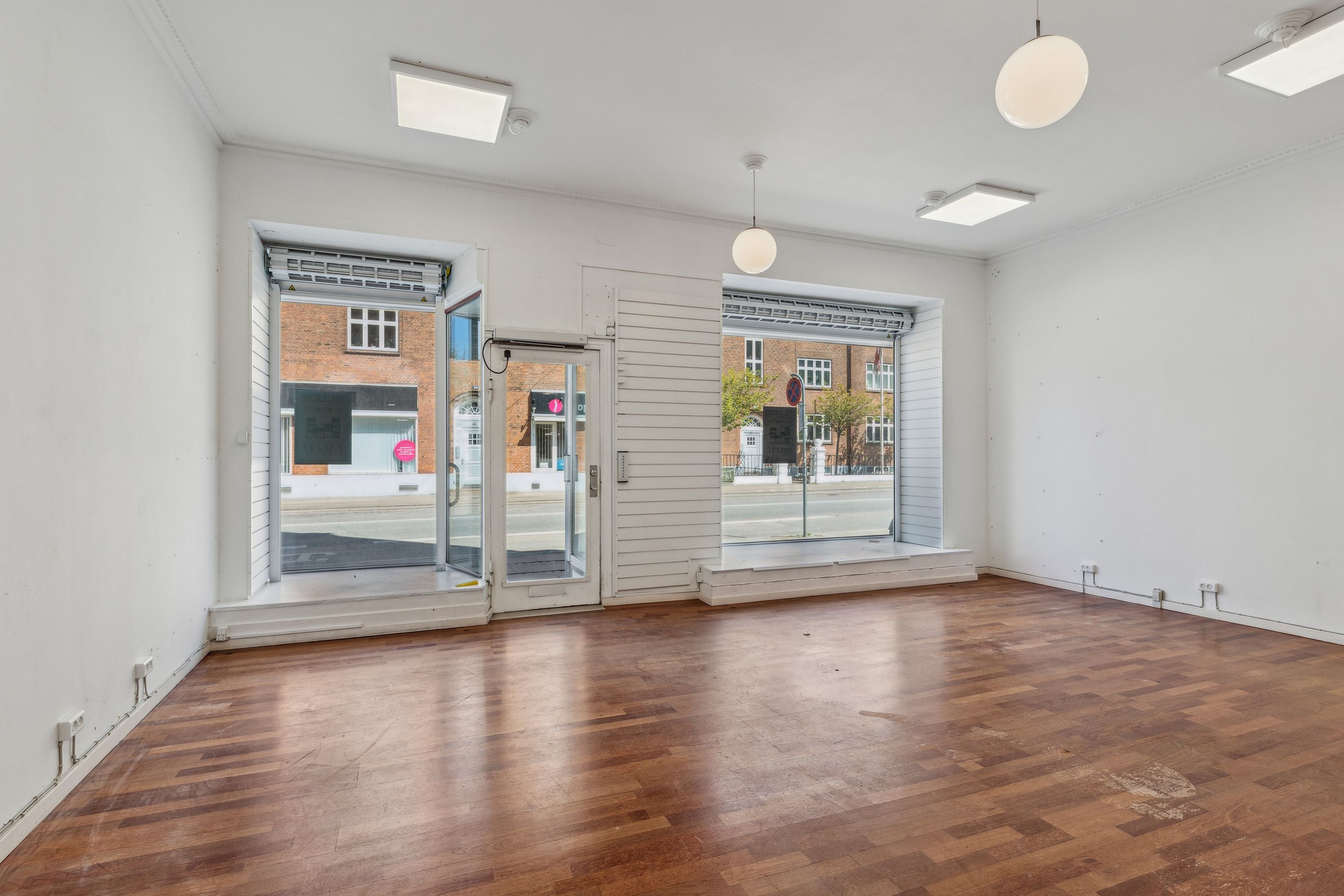 Foto af et tomt og lyset lokale med hårdt trægulv og store vinduer, hvorfra der er udsigt til en bygning og gade