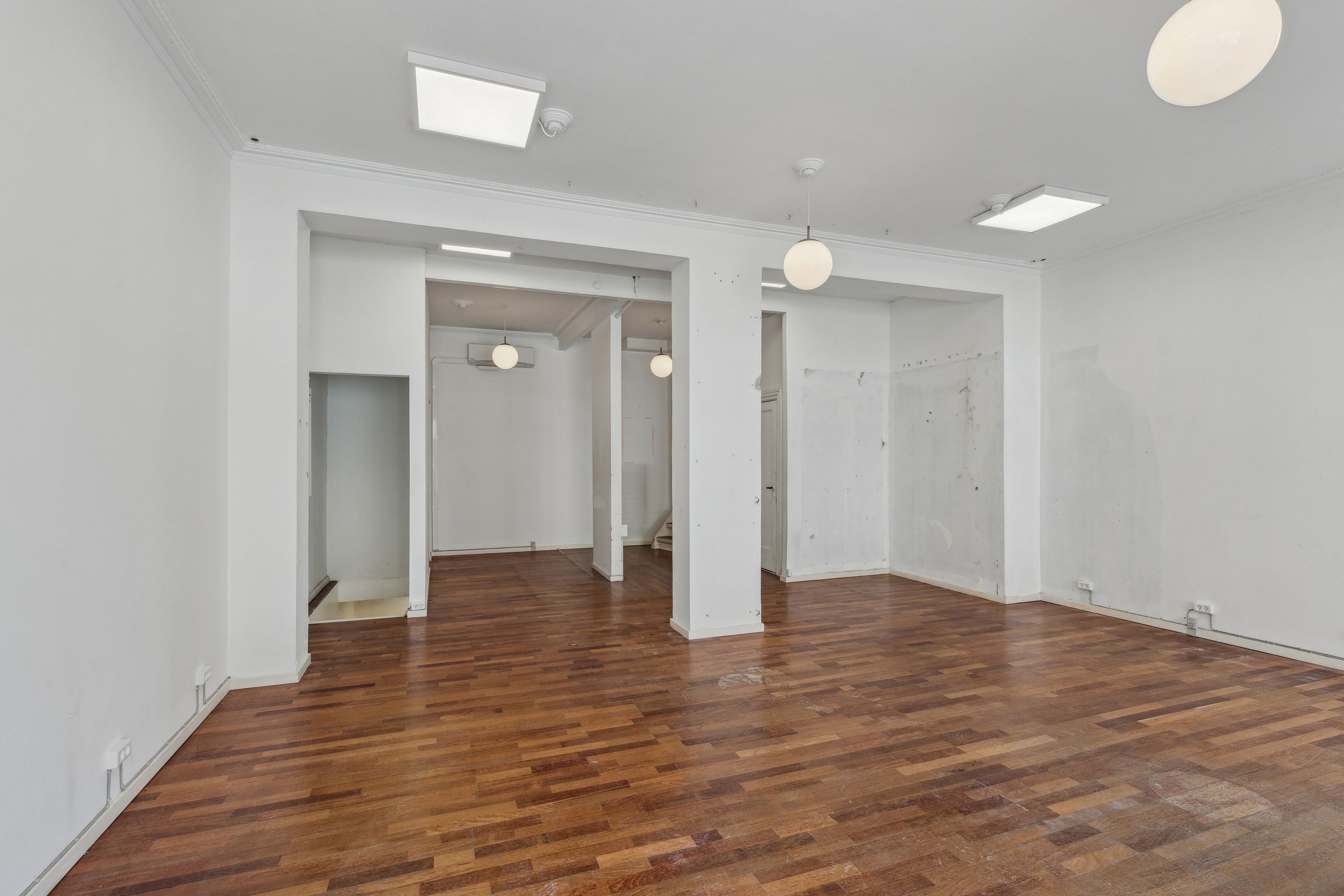 Et tomt rum med trægulv og hvide vægge, fire pendullampe og to lysekroner, samt en luftkonditioneringsventil i loftet.