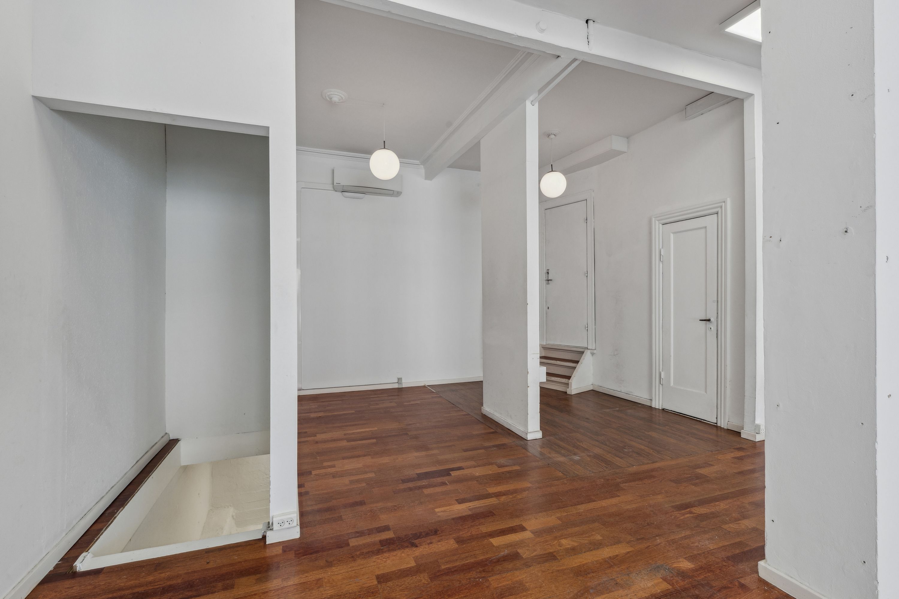 Et ubeboet rum med hårdt trægulv, to hvide vægge og to hvide døre, samt to hængende lamper og et klimaanlæg i loftet.