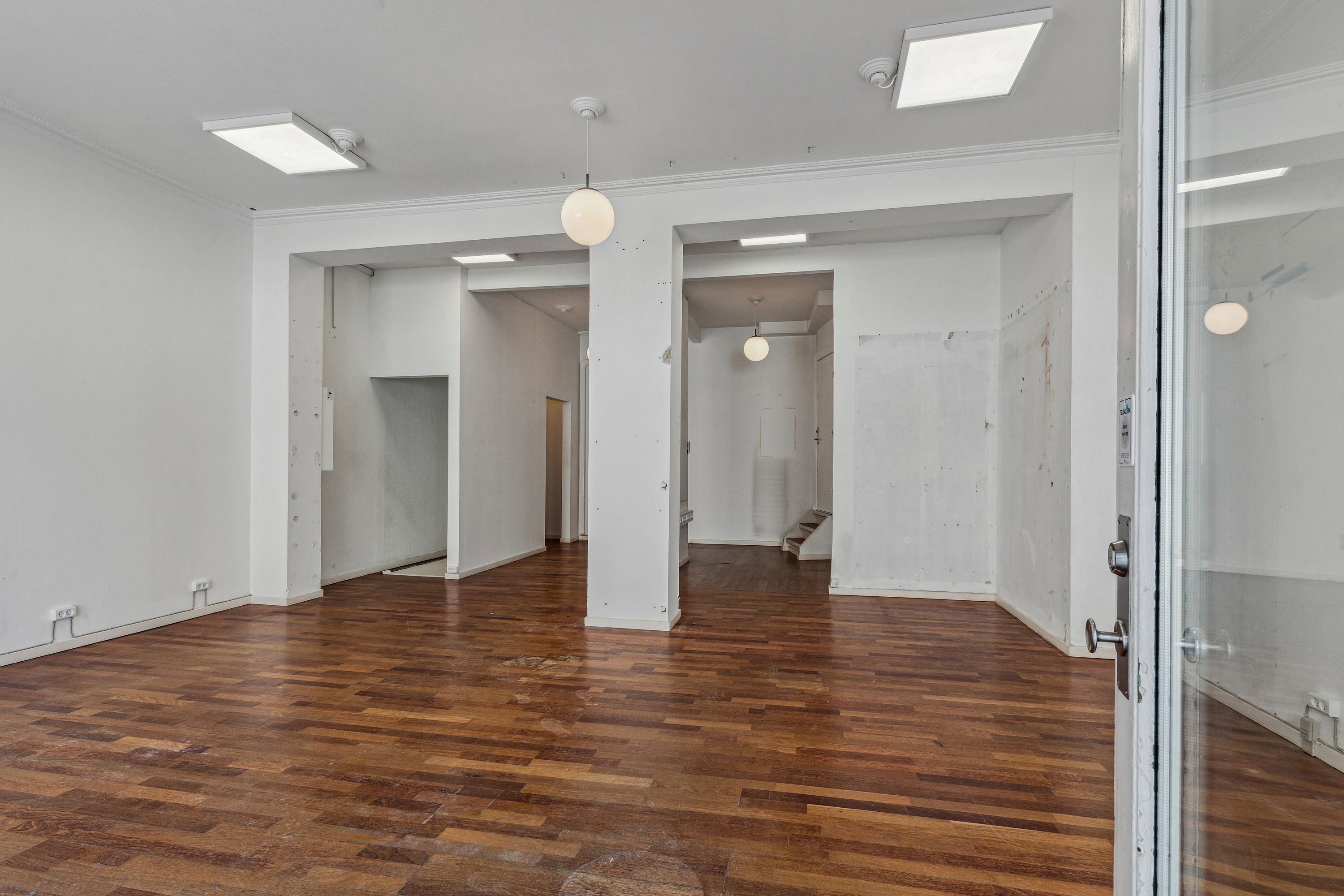 En stor stue med hårdt trægulv og hvide vægge. Der er åbne døre til siderum og lyskroner hængende fra loftet.